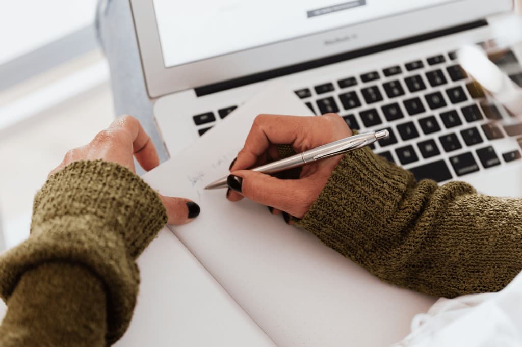 Hands writing in a notebook beside an open laptop